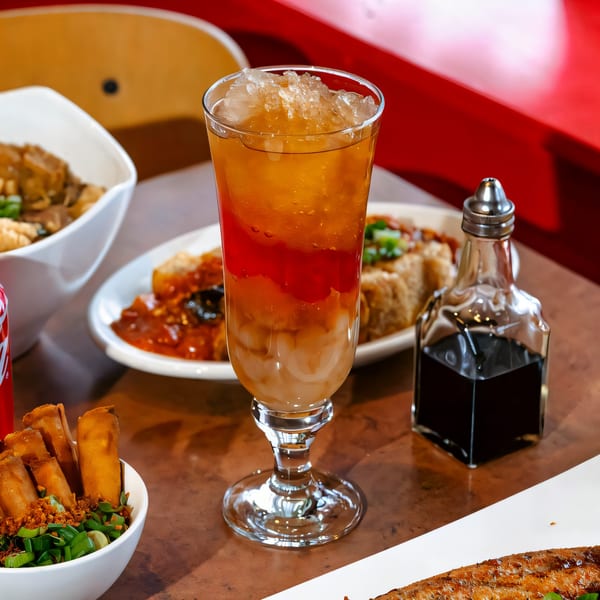 A tall glass of Filipino halo-halo with various colorful layers and crushed ice, surrounded by dishes.