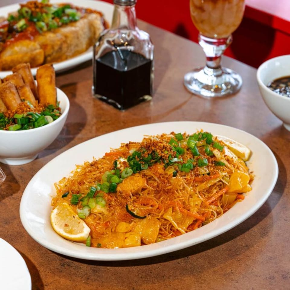 A plate of Filipino pancit bihon noodles with lemon, lumpia, and a drink on a table.