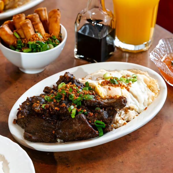 Filipino beef tapa with fried eggs and rice, alongside lumpia and orange juice.